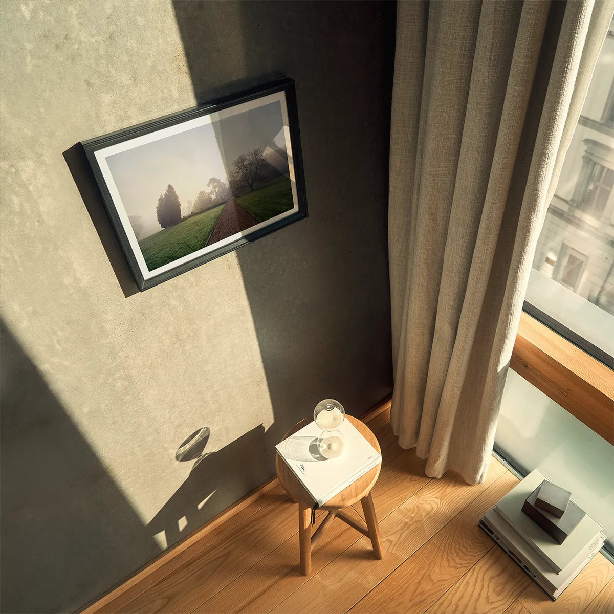 The image shows an interior space with a framed photograph of a foggy landscape hanging on a wall. The photo captures a misty morning in Surrey, England, with trees and a path visible. Sunlight streams through a window with beige curtains, casting shadows on the wall and floor. A wooden stool with a book and a glass sphere is placed near the window, adding a touch of elegance to the scene.